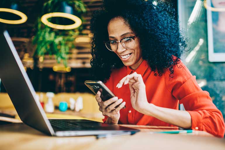 Vrouw bezig met smartphone, achter haar laptop.