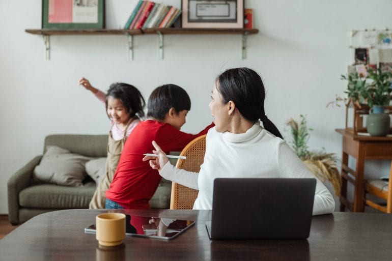 Foto van een vrouw met een laptop, met op de achtergrond spelende kinderen die de vrouw tot stilte maant