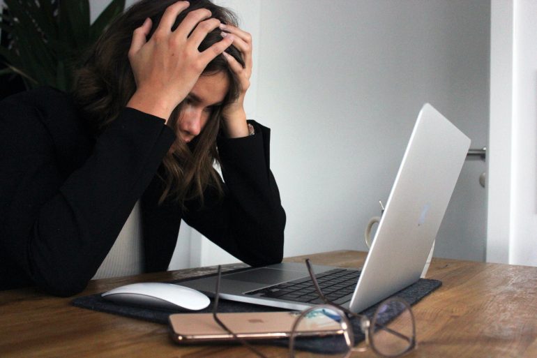Foto van een duidelijk gestresste persoon met een laptop, muis, telefoon en bril op het bureau.