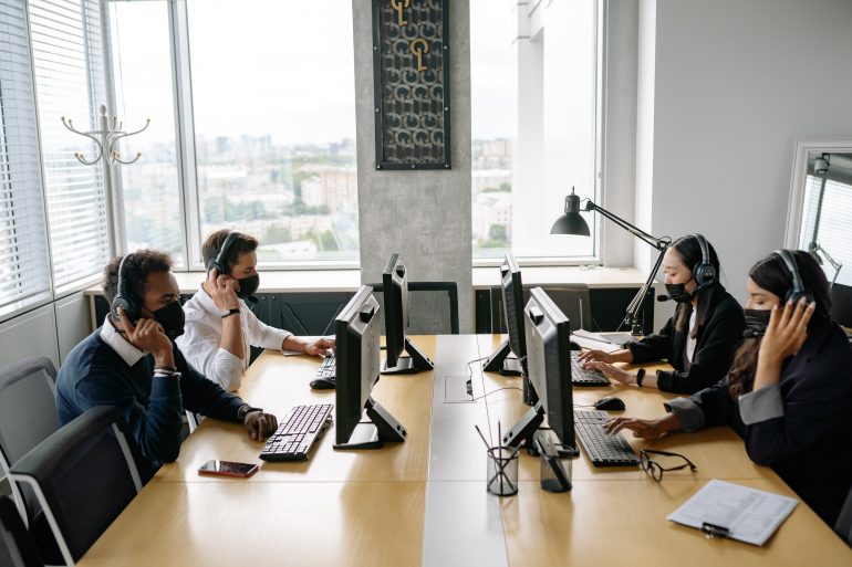 Groep mensen aan bureaus met koptelefoons en mondkapjes op.