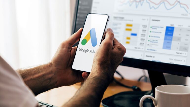 Een persoon houdt een telefoon vast met Google ads en data op zijn computer in de achtergrond.