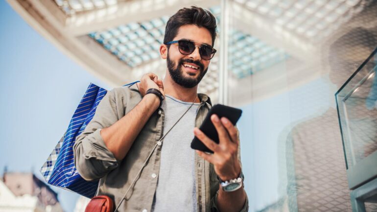 Man met winkeltassen kijkt op telefoon ter illustratie van mobile commerce