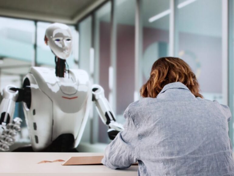 Een vrouw en robot tegenover elkaar aan tafel. De vrouw met de handen in het haar. 