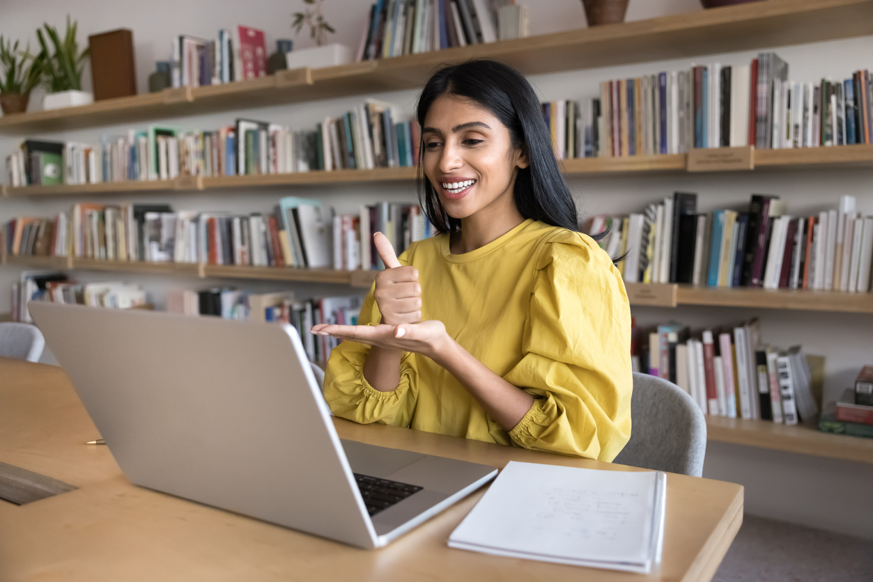 Sign language therapist talking on video call. Happy young Indian teacher woman speaking to student with hearing disability, showing hand gesture at laptop, working at computer in library